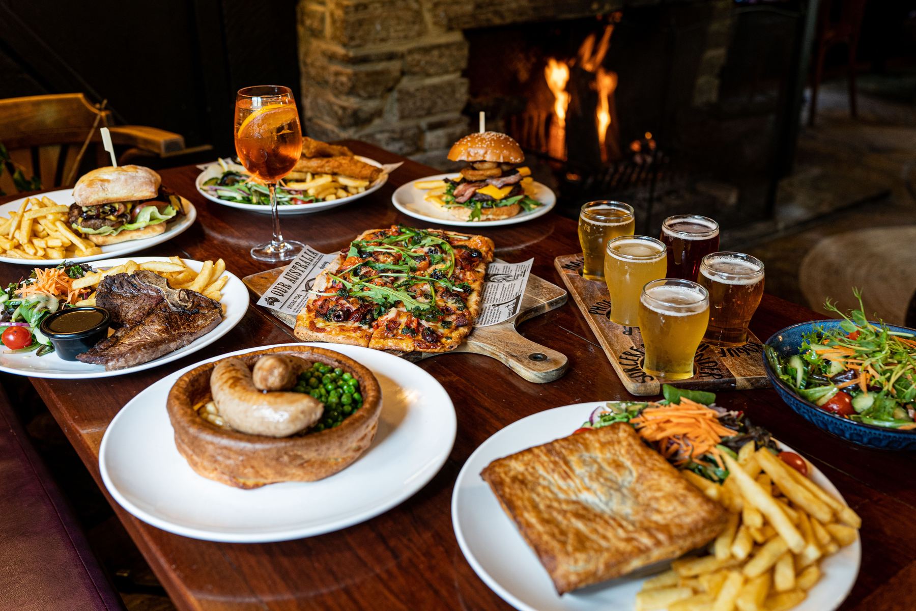 A spread of pub dishes and drinks — burgers, pizza, sausages and a beer flight — served in a traditional English pub.